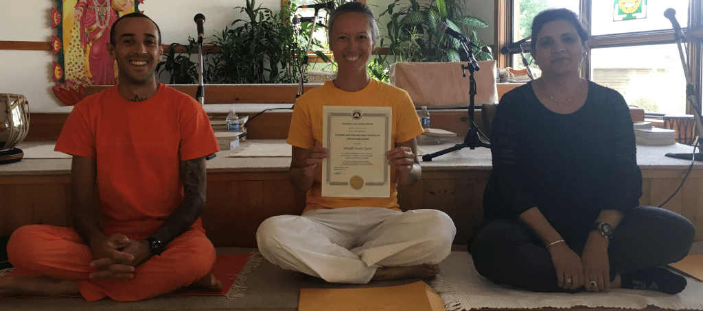 Jennifer holding training certificate. To her right is the teacher who gave the training Dr. Bhatt, and to her left is Swami Shivabhaktananda, the current director of the Yoga Camp.